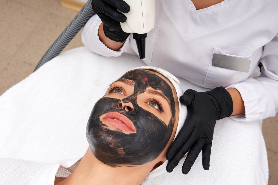 Cosmetologist applying black mask on the face of a beautiful woman for carbon peel